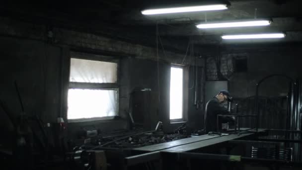 Un om într-un boilersuit negru în ochelari și un capac lustruiește o structură metalică a unui instrument de măcinat cu scântei pe masa atelierului sub lămpi de lumină naturală — Videoclip de stoc