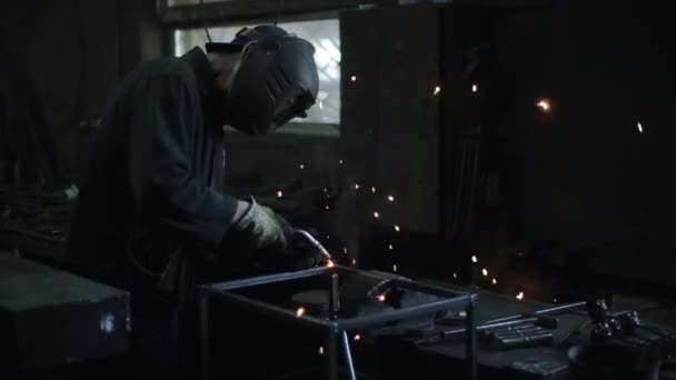 El cerrajero en un casco de soldadura y ropa especial sujeta una estructura metálica en la mesa del taller. Vista lateral — Vídeos de Stock