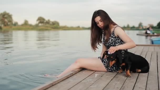 Красивая молодая девушка сидит на пирсе у реки и обнимает и гладит свою собаку таксу. Вид сбоку. Крупный план — стоковое видео
