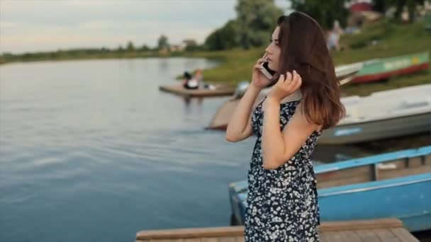 Chica joven agradable hablando en el teléfono móvil de pie en un muelle de madera junto al río. Vista desde un lado. Primer plano — Vídeos de Stock