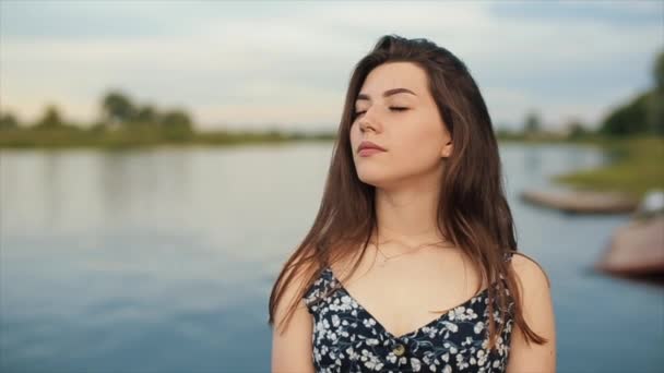 Retrato de una jovencita sensual que medita cerrando los ojos de pie en la orilla del río en un día de verano. Movimiento lento — Vídeos de Stock