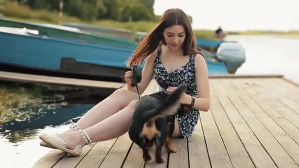 Довгошерста мила дівчина брюнетка грає зі своєю собакою такса, сидячи на дерев'яному пірсі біля річки в літній день. Крупним планом — стокове відео