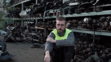 Gözlüklü ve gri tulumlu genç sakallı bir tamirci bir oto tamirhanesinin yedek parça deposunda oturup sigara içiyor. Yakın plan.