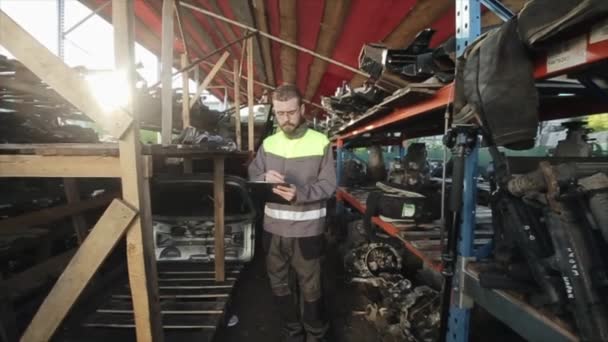 A young bearded auto mechanic with glasses walks between the shelves with a spare parts in the workshop and takes notes. View from the back. The camera follows him — Stock Video