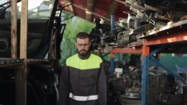Üniformalı, sakallı, ciddi bir tamirci kolları göğsünün üzerinde yedek parçalarla arka planda duruyor. Yakın plan.