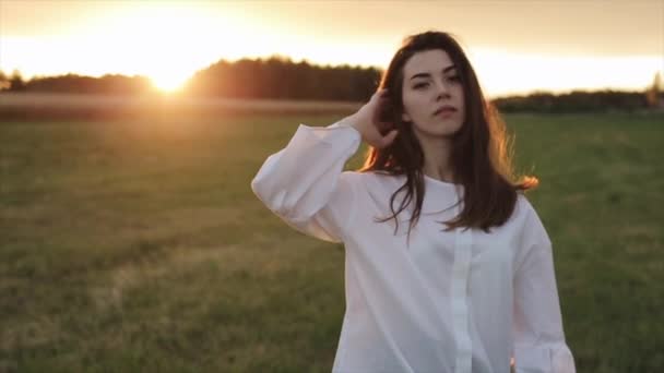 Una hermosa jovencita camina a través del campo en los rayos del atardecer y endereza su cabello con su mano. Primer plano. Movimiento lento — Vídeo de stock