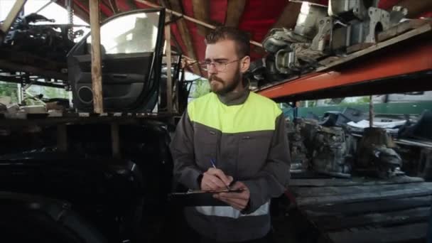 Mladý vousatý automechanik s brýlemi se prochází mezi policemi s náhradními díly a dělá si poznámky do zápisníku. Zastaví se v uličce a těžce vzdychne. Detailní záběr — Stock video