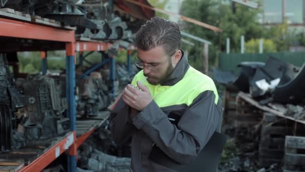 Un joven mecánico barbudo servicio de coches en un mono gris y gafas se sienta en una rotura de humo en el telón de fondo de los estantes con piezas de repuesto. Vista lateral. Primer plano — Vídeo de stock