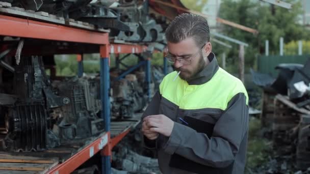 Un mecánico joven y cansado se tomó un descanso del trabajo y fumó mientras estaba sentado cerca de los bastidores con piezas de repuesto. Primer plano — Vídeo de stock