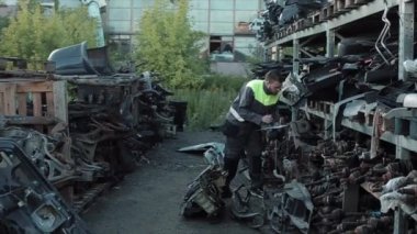 Gri tulumlu genç bir oto tamircisi yedek parça raflarıyla tepe örtüsünün altında yürür ve bir deftere notlar alır.