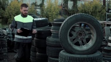 Genç sakallı araba tamircisi araba servisi deposunda araba tekerleğinin yanında duruyor. Yakın plan.