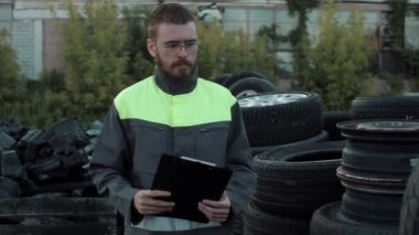 Genç sakallı bir tamirci üniformalı ve elinde bir defterle bir tamirhanenin bahçesinde kameraya bir şeyler söylüyor. Yakın plan.