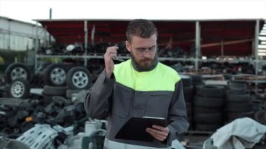 Gözlüklü genç tamirci araba servisinin yedek parçaları arasında duruyor kulağının arkasından kalem alıyor ve not defterine not alıyor. Yakın plan.