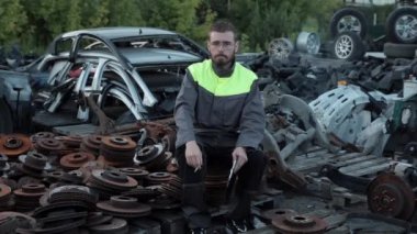 Gözlüklü genç sakallı tamirci bahçedeki katlanmış oto tamir parçalarının üzerinde otururken elini kameraya sallıyor.