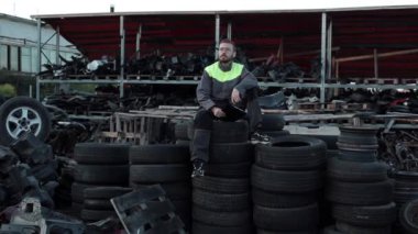 Gözlüklü genç sakallı araba tamircisi elinde telefon ve defterle katlanmış lastik yığınının üzerine oturur ve hayal kurarak gökyüzüne bakar.