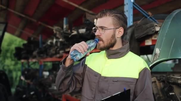 Un joven mecánico barbudo atractivo en gafas se encuentra en un almacén de piezas de automóviles y bebe agua de una botella. Primer plano — Vídeos de Stock