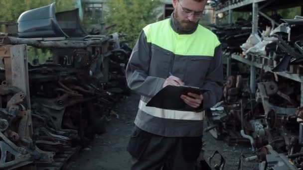 Un joven mecánico barbudo con gafas se encuentra entre los estantes con piezas de repuesto haciendo notas en un cuaderno y rascándose la cabeza pensativamente. Primer plano — Vídeos de Stock