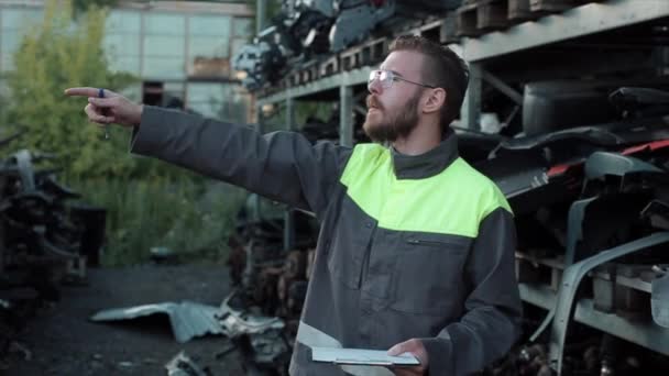 A young bearded mechanic with glasses stands near the racks with spare parts in a car service and gives instructions to someone. Close-up — Stock Video