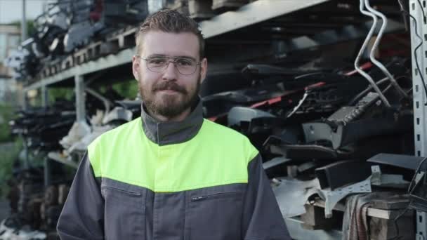 A young bearded auto mechanic in glasses stands against the background of a rack with spare parts and fooling around on camera making various grimaces. Close-up — Stock Video