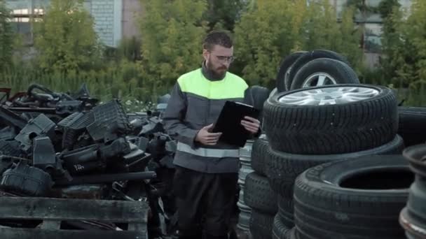 Un joven mecánico barbudo en uniforme comprueba neumáticos apilados en filas en el patio de un servicio de automóviles. Primer plano — Vídeos de Stock