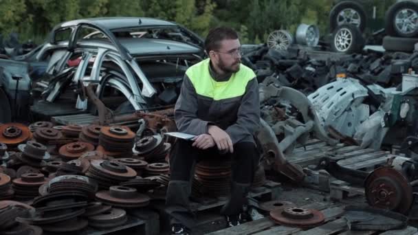 Tired young bearded mechanic with glasses sits on a pile of old disks among pallets with spare parts in the yard of a car workshop — Stock Video