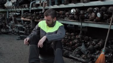 Genç, sakallı, gözlüklü ve üniformalı bir tamirci yedek parçalarla rafların yanında otururken sigara içiyor. Yakın plan.