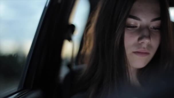 Una joven está hablando por video comunicación mientras está sentada en la ventana de un auto conduciendo a través de la ciudad nocturna. Primer plano — Vídeo de stock