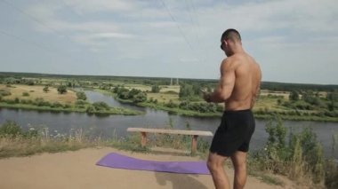 Çıplak gövdeli genç kaslı bir adam nehir kenarındaki bir tepede elinde telefonla duruyor ve bir spor paspası ayarlıyor. Yan görüş. Yakın plan.