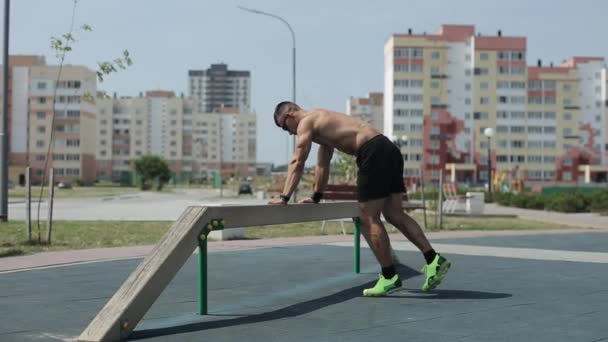 Giovane uomo muscoloso con torso nudo esegue esercizi sul terreno sportivo della città e non palmare sul fascio sportivo. Vista laterale — Video Stock