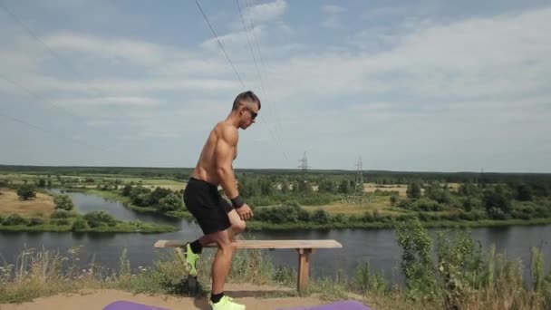 Çıplak gövdeli ve güneş gözlüğü takan kaslı bir adam nehir kıyısındaki bankta egzersiz yapıyor. Yan görüş. Kamera sporcuyu takip ediyor. — Stok video