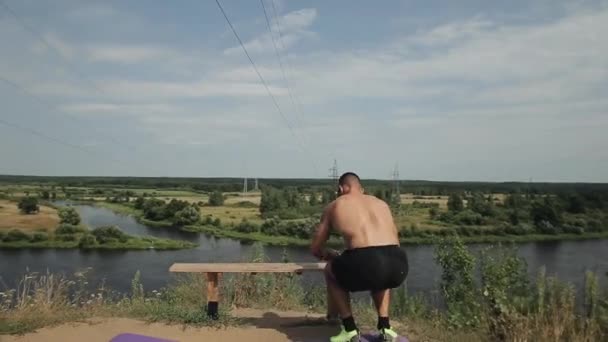 Siyah spor şortlu kaslı bir atlet nehir kıyısındaki bir bankta zıplayarak bacak egzersizi yapıyor. Arkadan bak. Yakın plan. — Stok video