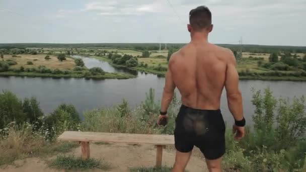 Young muscular man with a naked torso stands on a hill by the river and rests after training. View from the back. Close-up — Stock Video