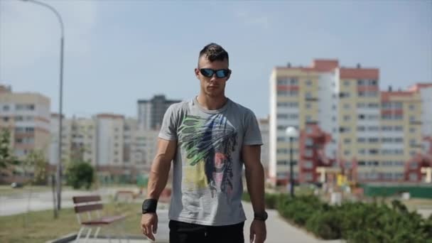 Misk, Belarus - 2017 년 5 월 15 일: 한 젊은 스포츠인이 창의적 인 티셔츠와 선글라스를 끼고 여름 도시를 걷고 있다. 앞을 봐. 클로즈업. 느린 동작. 실명 한 백골 족 — 비디오