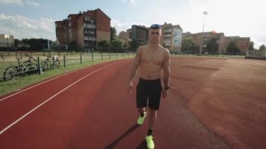 Spor şortlu ve parlak spor ayakkabılı çıplak gövdeli genç kaslı bir adam stadyumun koşu bandında yürüyor. Yakın plan. Kamera yukarı ve aşağı hareket eder