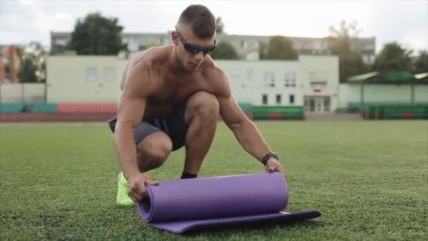 A muscular man with a naked torso and sunglasses rolls up the sports mat after training at the city stadium. Close-up. Slow motion — Stock Video