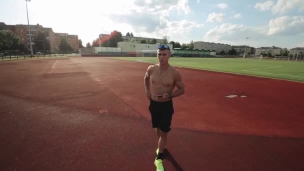 A young muscular man with a naked torso walks through the city stadium with a phone in hand and speaks to the camera. The camera zooms in and out — Stock Video