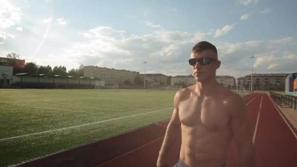 En ung sportig man i solglasögon och en naken bål står på stadens stadion mot bakgrund av den soliga himlen. Närbild — Stockvideo