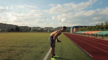 Çıplak gövdeli genç kaslı adam stadyumda egzersiz yaptıktan sonra bacak kaslarını ovalıyor. Kamera konunun etrafında hareket ediyor.