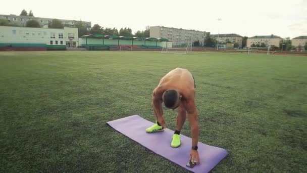 Un tipo musculoso con un torso desnudo en un estadio de la ciudad hace ejercicios de piernas con un temporizador en su teléfono móvil. Estilo de vida saludable. Primer plano — Vídeos de Stock