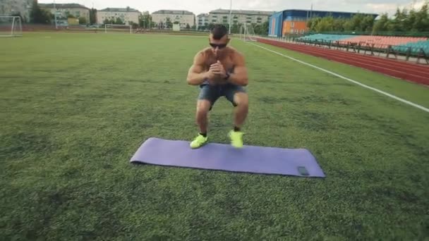 Ένας bodybuilder με αθλητικά σορτς και γυαλιά ηλίου κάνει ασκήσεις στο γήπεδο ποδοσφαίρου του σταδίου της πόλης με χρονόμετρο στο κινητό του τηλέφωνο.. Η κάμερα κινείται πίσω από τον αθλητή — Αρχείο Βίντεο