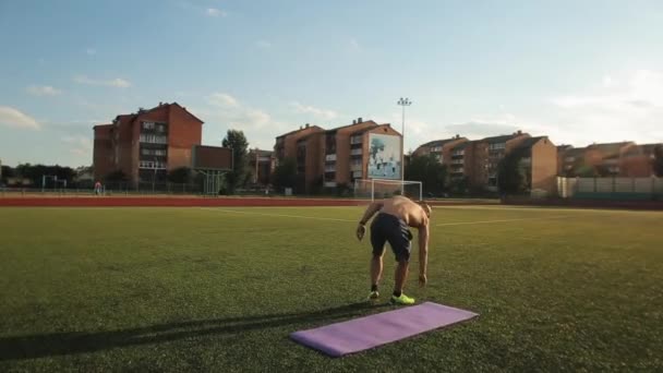 Un joven musculoso con un torso desnudo lleva una alfombra deportiva en el estadio de la ciudad para entrenar. Estilo de vida saludable. Vista trasera — Vídeos de Stock