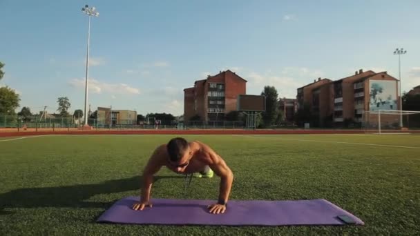 Hombre musculoso con un torso desnudo en gafas de sol haciendo flexiones en una alfombra deportiva en el estadio de la ciudad. Primer plano. Vista frontal. La cámara sigue los movimientos. Sportsman utiliza la aplicación móvil en su teléfono para — Vídeos de Stock