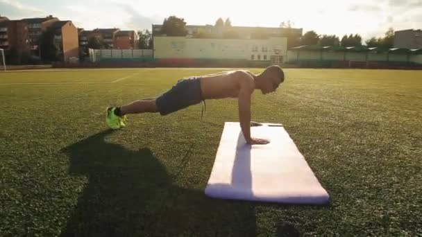 Sportsman utiliza la aplicación móvil de su entrenamiento telefónico. Un hombre bombeado entrenando en un estadio de la ciudad y hace flexiones moviendo sus manos sobre la alfombra. Vista lateral. Primer plano — Vídeos de Stock