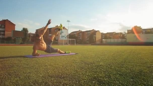 裸の胴を持つ筋肉質の男がスタジアムでトレーニングし、横板に立っている間に足をスイングします。接近中だ。カメラが前に戻る — ストック動画