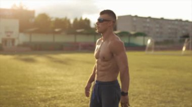 Spor şortlu bir vücut geliştirici stadyumda antrenman yapıyor ve gün batımında bacaklarını sallıyor. Sağlıklı bir yaşam tarzı. Yan görüş. Yakın plan.