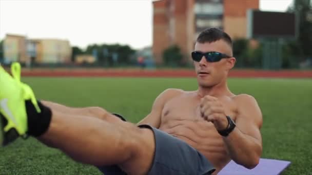 An athletic man with a naked torso and in sunglasses doing abs exercises while lying on a mat in a city stadium. Close-up. Front view. Blurred background — Stock Video