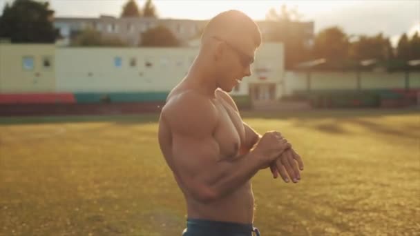 Hombre musculoso torso desnudo recuperándose de entrenar en el estadio sacudiendo las manos y mira su reloj. Primer plano. Fondo borroso — Vídeos de Stock