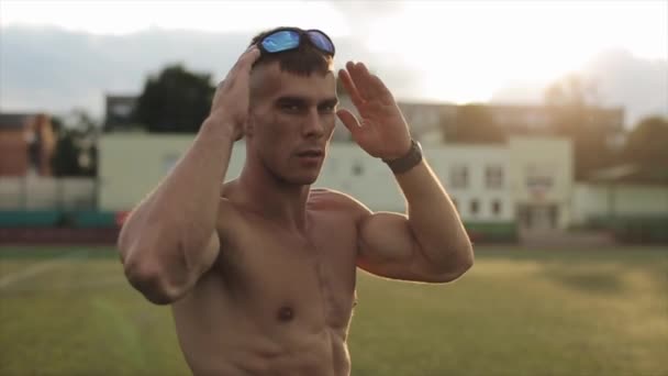 Muskularny mężczyzna z nagim tułowiem po treningu stoi na stadionie i zakłada okulary przeciwsłoneczne. Z przodu. Zbliżenie. Zamazane tło — Wideo stockowe