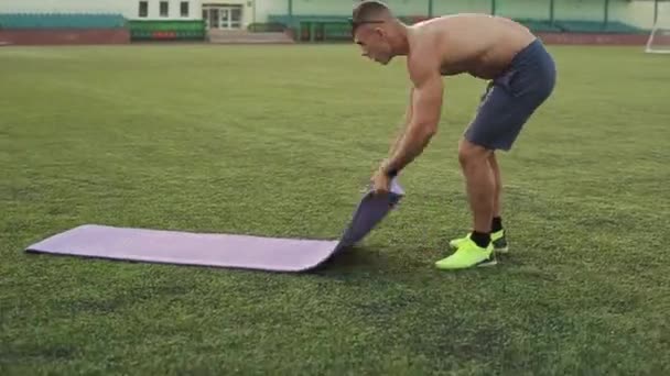 Hombre atlético en pantalones cortos azules pliega la alfombra deportiva después de entrenar en el estadio de la ciudad. Vista lateral. Primer plano — Vídeo de stock