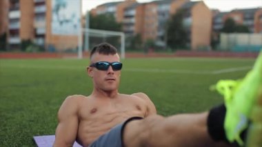 Çıplak gövdeli ve güneş gözlüklü bir sporcu, stadyumdaki bir mindere uzanmış karın kası egzersizi yapıyor. Yakın plan. Ön manzara. Bulanık arkaplan